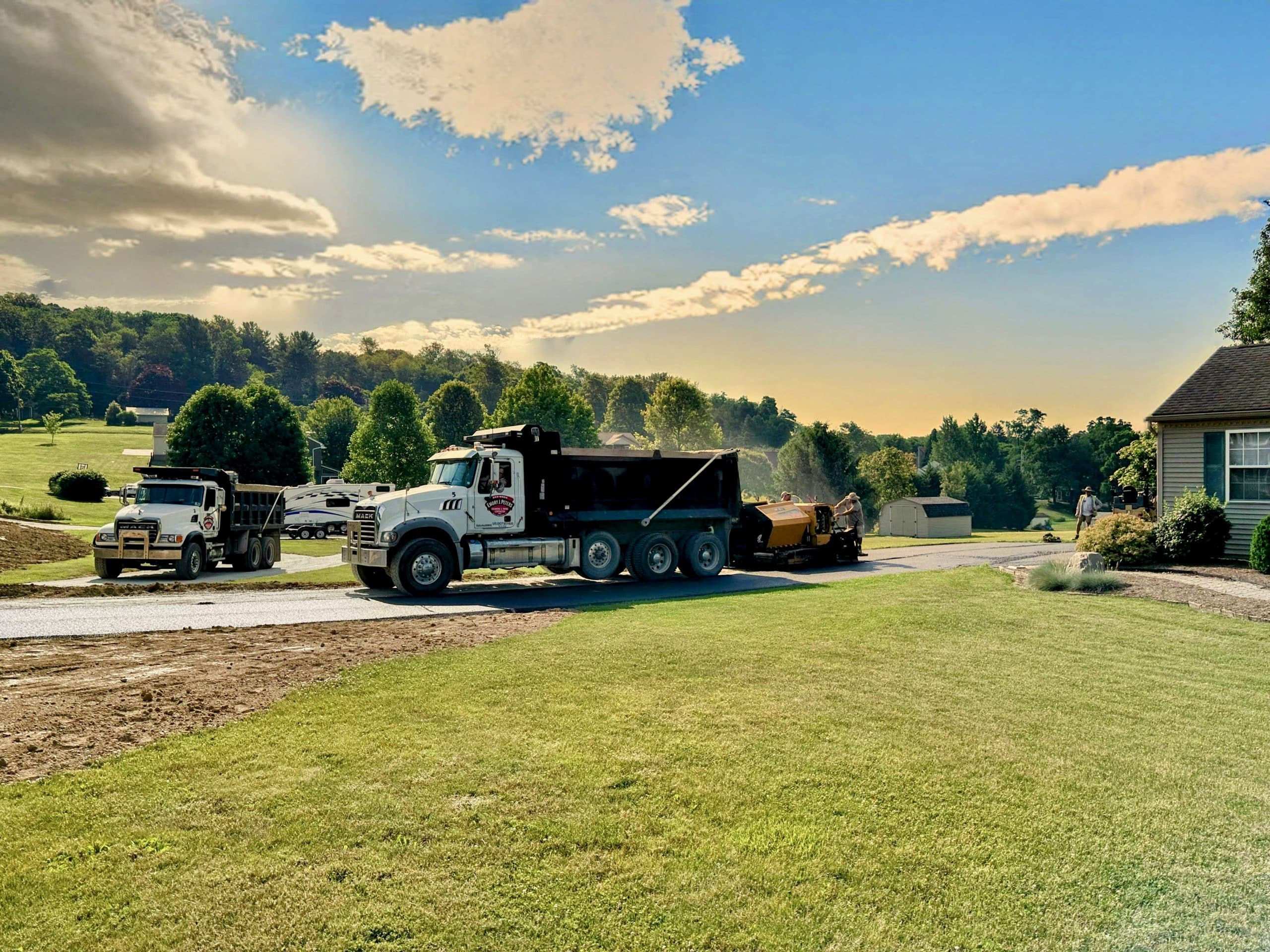 A picturesque construction site with multiple trucks parked on a paved road bordered by lush green fields and trees, creating the perfect natural footer to the scene. The sky is clear with a few clouds, while workers are visible in the distance.