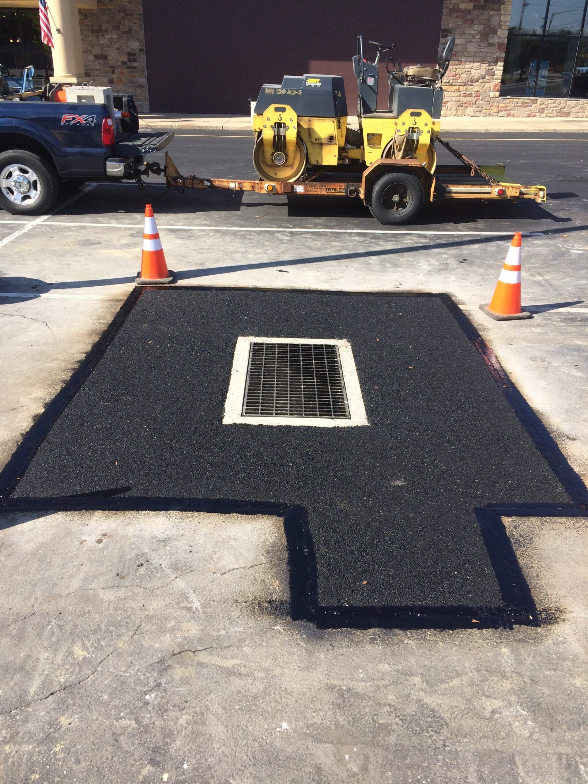 A newly paved section of asphalt with a rectangular drainage grate in the center is surrounded by orange traffic cones. A truck and a yellow asphalt roller are parked nearby.